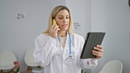Téléchargez les photos : Jeune femme blonde médecin en utilisant touchpad parler sur smartphone à la salle d'attente de la clinique - en image libre de droit