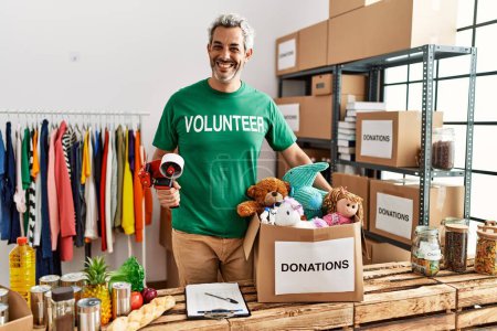 Foto de Middle age grey-haired man volunteer smiling confident holding packing machine at charity center - Imagen libre de derechos