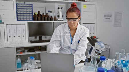 Téléchargez les photos : Portrait d'une jeune scientifique rousse concentrée, profondément engagée dans sa recherche en biologie sur un ordinateur portable à l'intérieur du laboratoire, démêlant les mystères médicaux de la science avec une expression sérieuse. - en image libre de droit