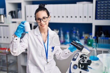 Photo for Young caucasian woman scientist using microscope holding pills bottle at laboratory - Royalty Free Image