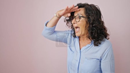 Foto de Mujer hispana de mediana edad buscando a alguien sorprendido sobre un fondo rosa aislado - Imagen libre de derechos
