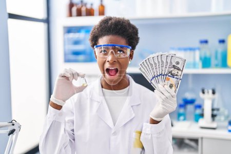 Foto de Mujer afroamericana trabajando en laboratorio científico sosteniendo dólares enojada y loca gritando frustrada y furiosa, gritando con ira mirando hacia arriba. - Imagen libre de derechos