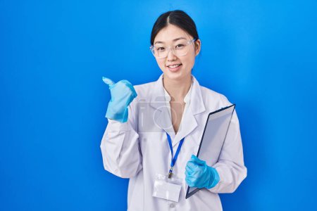 Téléchargez les photos : Jeune femme chinoise travaillant au laboratoire scientifique pointant le dos derrière avec la main et les pouces levés, souriant confiant - en image libre de droit