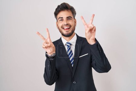 Photo for Young hispanic man with tattoos wearing business suit and tie smiling with tongue out showing fingers of both hands doing victory sign. number two. - Royalty Free Image