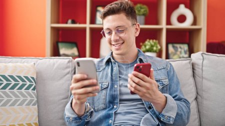 Photo for Young hispanic man using smartphones sitting on sofa at home - Royalty Free Image