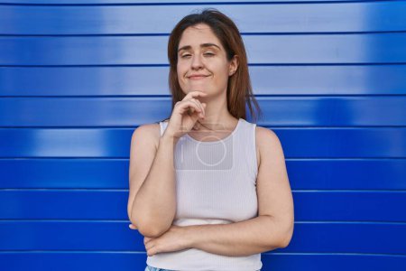 Photo for Brunette woman standing over blue background smiling looking confident at the camera with crossed arms and hand on chin. thinking positive. - Royalty Free Image