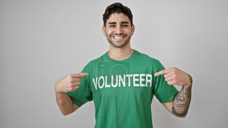 Téléchargez les photos : Jeune homme hispanique pointant vers uniforme volontaire souriant sur fond blanc isolé - en image libre de droit