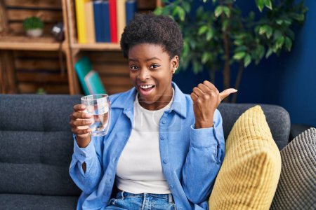 Foto de Mujer afroamericana bebiendo vaso de agua apuntando con el pulgar hacia el costado sonriendo feliz con la boca abierta - Imagen libre de derechos