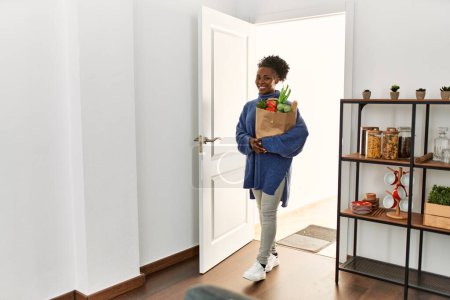 Téléchargez les photos : Femme afro-américaine tenant sac d'épicerie marchant à travers la porte à la maison - en image libre de droit