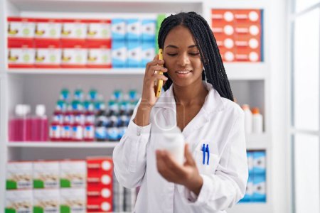Téléchargez les photos : Une pharmacienne afro-américaine parle sur un smartphone tenant des pilules à la pharmacie - en image libre de droit