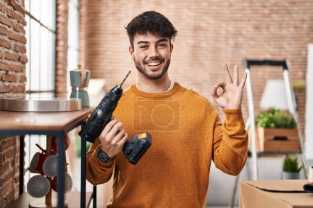 Foto de Hombre hispano con barba sosteniendo destornillador en casa nueva haciendo signo ok con los dedos, sonriendo gesto amistoso excelente símbolo - Imagen libre de derechos