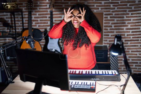 Téléchargez les photos : Plus taille femme hispanique jouant du piano au studio de musique faisant un geste correct comme des jumelles sortant la langue, les yeux regardant à travers les doigts. expression folle. - en image libre de droit