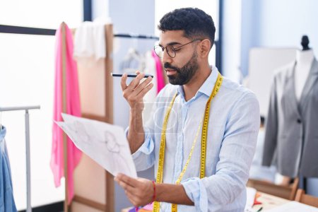 Téléchargez les photos : Jeune homme arabe tailleur parler sur smartphone conception de vêtements à la recherche au magasin de tailleur - en image libre de droit