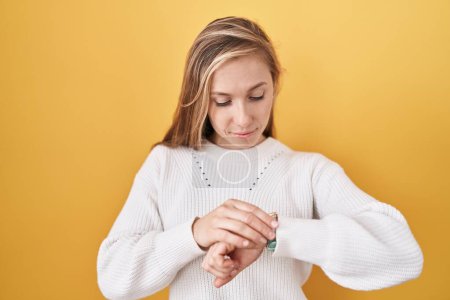 Téléchargez les photos : Jeune femme caucasienne portant un pull blanc sur fond jaune vérifiant l'heure sur la montre-bracelet, détendue et confiante - en image libre de droit