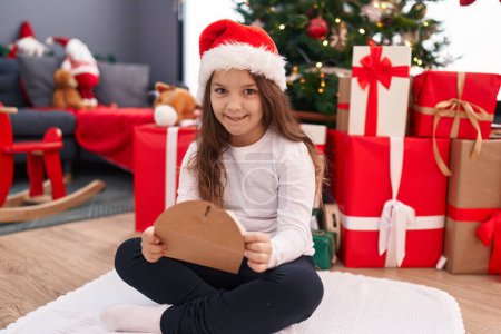 Photo for Adorable hispanic girl holding santa claus letter sitting on floor by christmas tree at home - Royalty Free Image