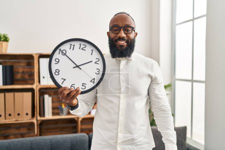Téléchargez les photos : Afro-Américain tenant grande horloge au bureau regardant positif et heureux debout et souriant avec un sourire confiant montrant les dents - en image libre de droit