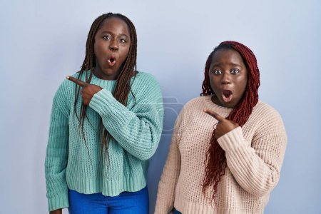 Téléchargez les photos : Deux femmes africaines debout sur fond bleu surpris pointant du doigt sur le côté, bouche ouverte expression étonnée. - en image libre de droit