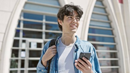 Téléchargez les photos : Jeune homme hispanique étudiant souriant confiant en utilisant un smartphone à l'université - en image libre de droit