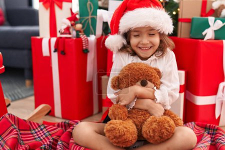 Foto de Adorable chica hispana abrazando oso de peluche sentado en el suelo por el árbol de Navidad en casa - Imagen libre de derechos