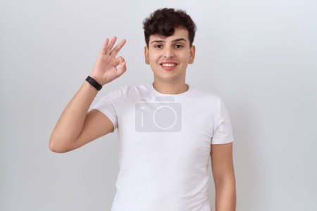 Foto de Joven hombre no binario vistiendo casual camiseta blanca sonriendo positiva haciendo signo de ok con la mano y los dedos. expresión exitosa. - Imagen libre de derechos