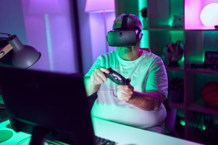 Foto de Streamer hombre de pelo gris de mediana edad jugando videojuegos con gafas de realidad virtual y joystick en la sala de juegos - Imagen libre de derechos