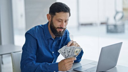 Téléchargez les photos : Jeune homme hispanique travailleur d'affaires en utilisant ordinateur portable comptant des dollars au bureau - en image libre de droit