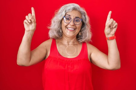 Photo for Middle age woman with grey hair standing over red background smiling amazed and surprised and pointing up with fingers and raised arms. - Royalty Free Image