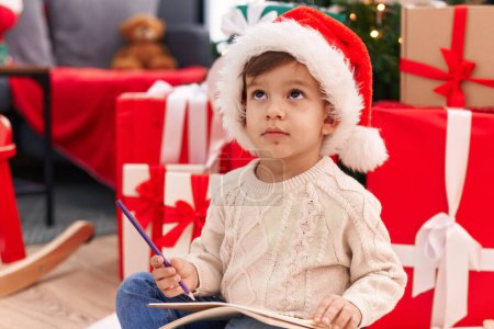 Foto de Adorable niño hispano escribiendo carta de Santa Claus sentado en el suelo por el árbol de Navidad en casa - Imagen libre de derechos