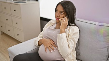 Photo for Emotional young pregnant woman, sitting at home on her sofa, talks and shares her doubts and ideas on her smartphone, touching her belly while in deep thought - Royalty Free Image