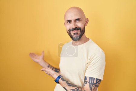Téléchargez les photos : Homme hispanique avec des tatouages debout sur fond jaune invitant à entrer souriant naturel avec la main ouverte - en image libre de droit