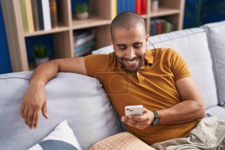 Photo for Young latin man using smartphone sitting on sofa at home - Royalty Free Image