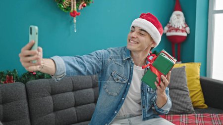 Foto de Joven hombre hispano tomando una foto selfie sosteniendo regalo de Navidad en casa - Imagen libre de derechos