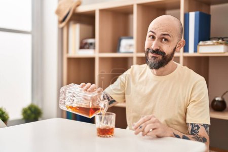 Foto de Young bald man pouring whisky on glass sitting on table at home - Imagen libre de derechos