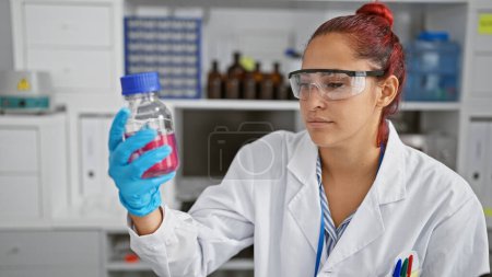 Foto de Intenso enfoque, hermosa, joven pelirroja irlandesa científica trabajando vigorosamente en el laboratorio, la medición experta de líquido en una botella con guantes de seguridad, en medio de la tecnología de investigación médica. - Imagen libre de derechos