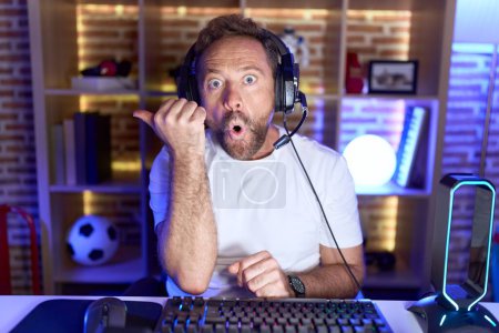 Foto de Hombre de mediana edad con barba jugando videojuegos con auriculares sorprendidos apuntando con el dedo de la mano hacia un lado, boca abierta expresión asombrada. - Imagen libre de derechos