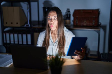 Photo for Young brunette woman working at the office at night making fish face with lips, crazy and comical gesture. funny expression. - Royalty Free Image