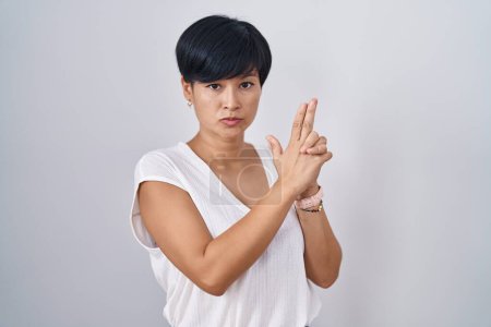 Foto de Joven mujer asiática con pelo corto de pie sobre fondo aislado sosteniendo arma simbólica con gesto de mano, jugando a matar armas de fuego, cara enojada - Imagen libre de derechos