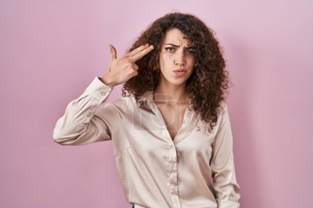 Foto de Mujer hispana con el pelo rizado de pie sobre fondo rosa disparando y matándose señalando la mano y los dedos a la cabeza como arma, gesto suicida. - Imagen libre de derechos
