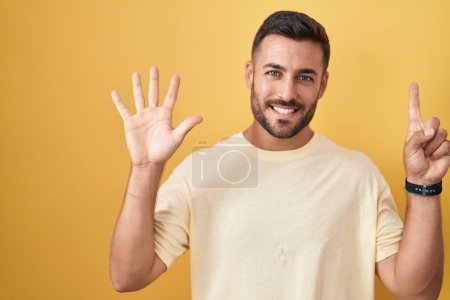 Foto de Hombre hispano guapo de pie sobre fondo amarillo mostrando y señalando hacia arriba con los dedos número seis mientras sonríe confiado y feliz. - Imagen libre de derechos