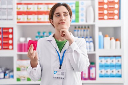 Téléchargez les photos : Femme brune travaillant à la pharmacie drugstore tenant préservatif visage sérieux penser à la question avec la main sur le menton, réfléchi sur l'idée déroutante - en image libre de droit