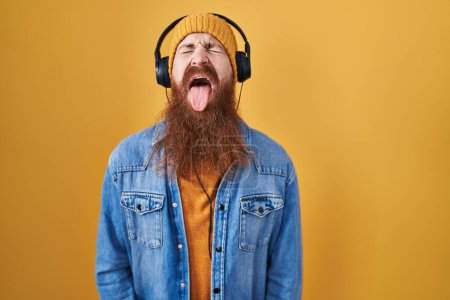 Téléchargez les photos : Homme caucasien à longue barbe écoutant de la musique à l'aide d'écouteurs qui sortent la langue heureuse avec une expression drôle. concept d'émotion. - en image libre de droit