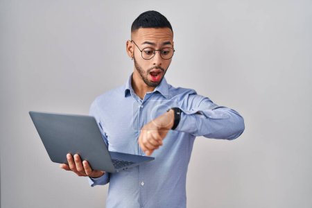 Téléchargez les photos : Jeune homme hispanique travaillant à l'aide d'un ordinateur portable en regardant le temps de la montre inquiet, peur de se faire tard - en image libre de droit