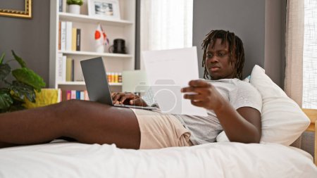 Photo for African american man using laptop reading document lying on bed at bedroom - Royalty Free Image