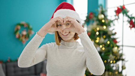 Photo for Young blonde woman smiling by christmas tree doing heart shape with hands at home - Royalty Free Image