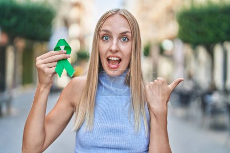 Téléchargez les photos : Jeune femme caucasienne tenant soutien ruban vert pointant pouce vers le côté souriant heureux avec bouche ouverte - en image libre de droit