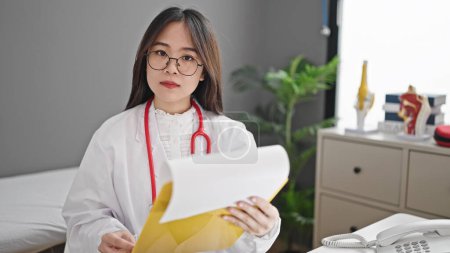 Photo for Young chinese woman doctor reading document on clipboard at clinic - Royalty Free Image