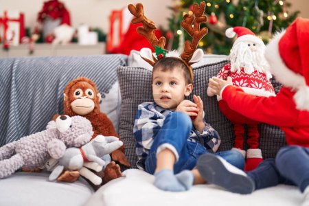 Photo for Adorable toddler wearing reindeer ears sitting on sofa by christmas tree at home - Royalty Free Image