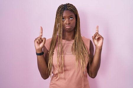 Photo for African american woman with braided hair standing over pink background pointing up looking sad and upset, indicating direction with fingers, unhappy and depressed. - Royalty Free Image