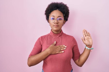 Foto de Hermosa mujer africana con el pelo rizado de pie sobre fondo rosa jurando con la mano en el pecho y la palma abierta, haciendo una promesa de lealtad juramento - Imagen libre de derechos