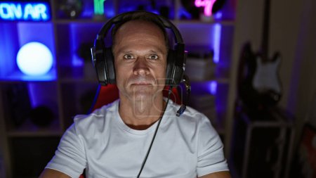 Photo for Serious-faced middle age man streamer, immersed in the digital world of gaming inside his home office - handsome hispanic gamer, rocking headphones, streaming cyber entertainment through the night - Royalty Free Image